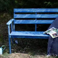 Niebieska ławka ogrodowa z kocem i czasopismem, obok konewka i kwiaty.