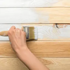 Dłoń maluje pędzlem drewniane deski na biało.