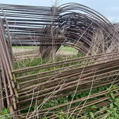 Stos zardzewiałych, metalowych pałąków do tunelu foliowego, otoczony dziką roślinnością.