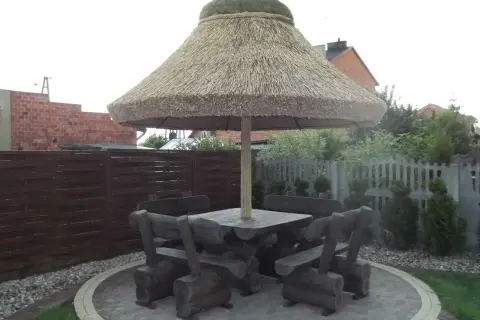Drewniany stół z ławami pod dużym, słomianym parasolem ogrodowym.