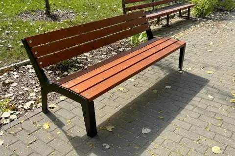 Drewniana ławka ogrodowa z czarnymi metalowymi nogami na wybrukowanym chodniku.