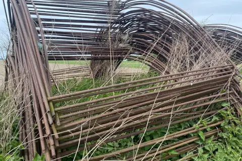 Stos zardzewiałych, metalowych pałąków do tunelu foliowego, otoczony dziką roślinnością.
