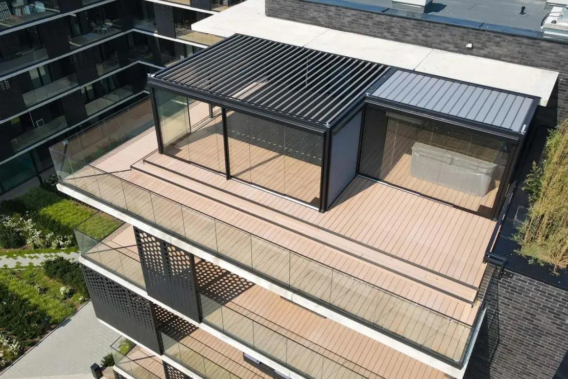 Nowoczesny taras na dachu z przeszklonym pawilonem i pergolą. Drewniana podłoga i szklane balustrady tworzą elegancką przestrzeń.