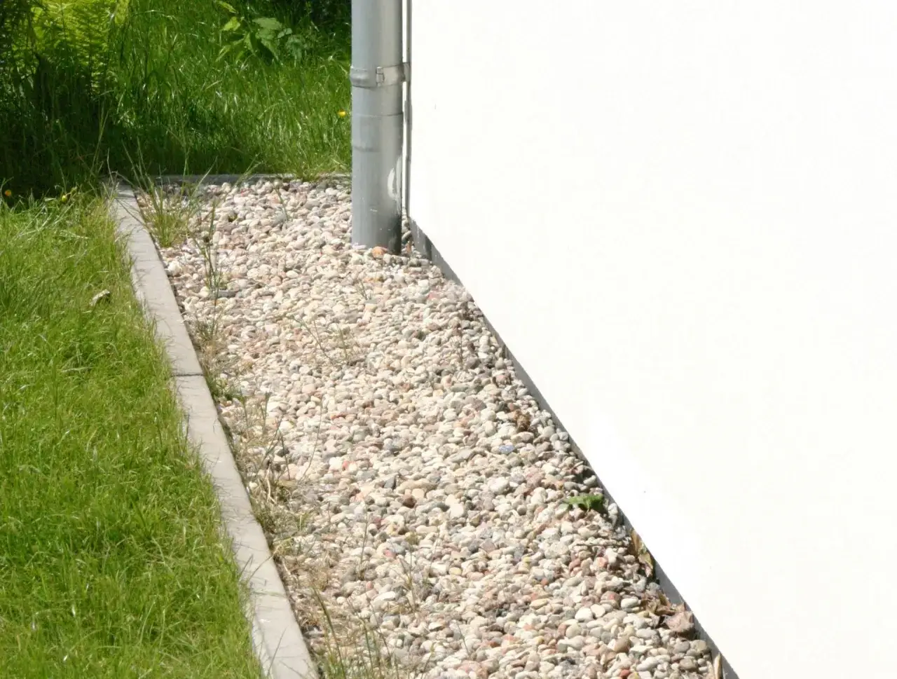 Opaska wokół domu z kamienia, oddzielona od trawnika betonowym krawężnikiem. Rynna odprowadza wodę od białej ściany.