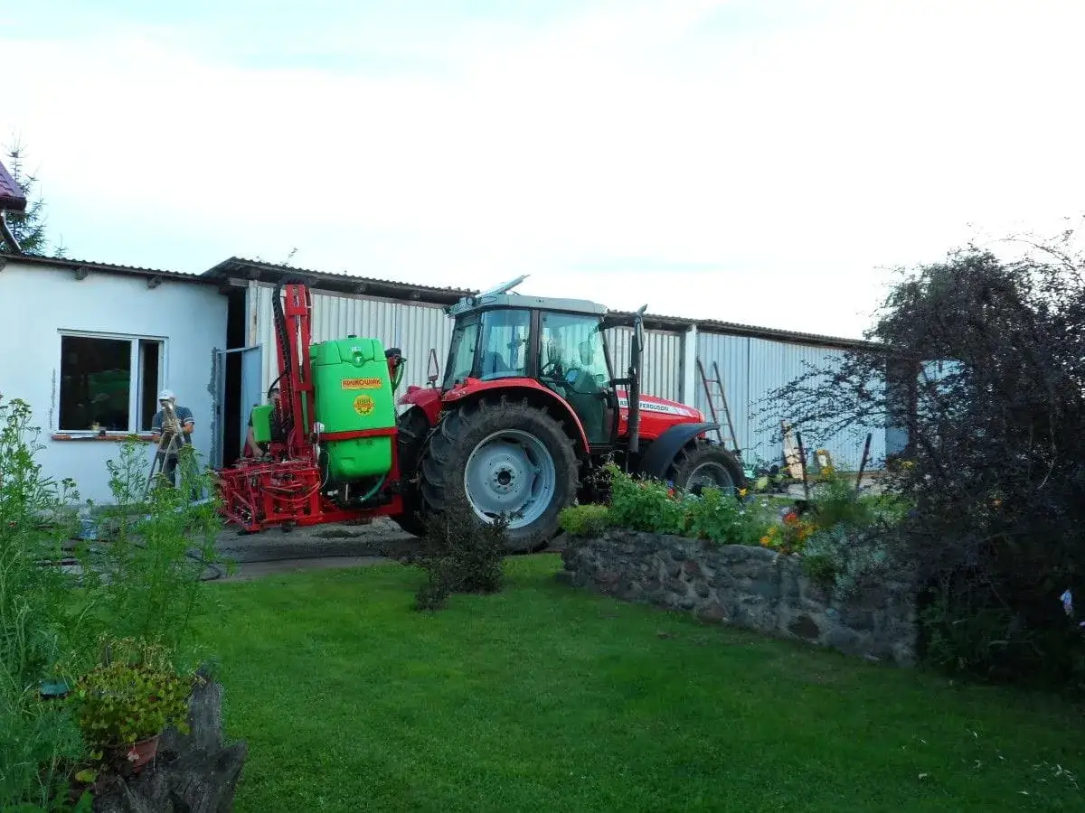 Czerwony traktor z opryskiwaczem czeka na deszcz. Rolnik na drabinie przy budynku.