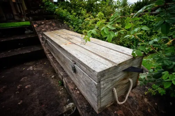 Drewniana skrzynia ogrodowa z linowymi uchwytami, na kamiennym murku, otoczona zielenią.